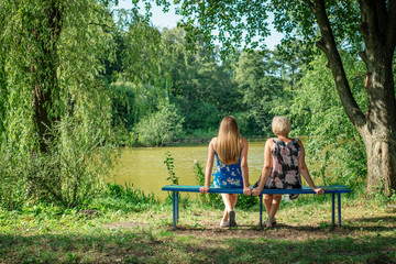 Two women of different generations sitting on a bench near a pond in the summer. Mother and daughter hugging. Grandmother and granddaughter.