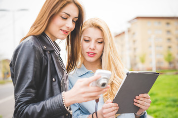 Two young blonde and brunette girls walking through the city using a tablet and a camera -...