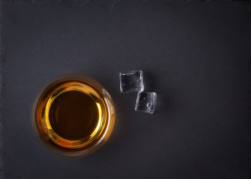 Glass of whiskey with ice on black stone table. Top view with copy space