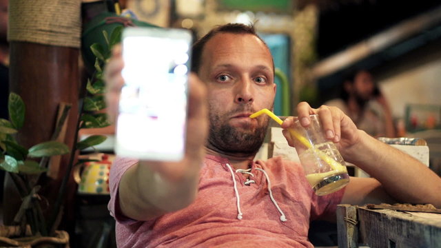 Young man taking selfie photo with cellphone and drinking cocktail in cafe 
