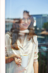 Young woman standing behind glass of balcony door, drinking morning coffee