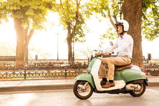 Trendy Man Driving A Scooter In Helmet. Sun Is Shining Through Trees On Background