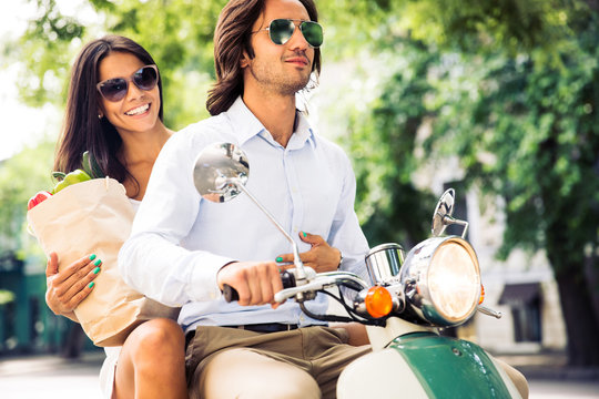 Fototapeta Happy young couple driving scooter while woman holding bag full of groceries. Summer time.