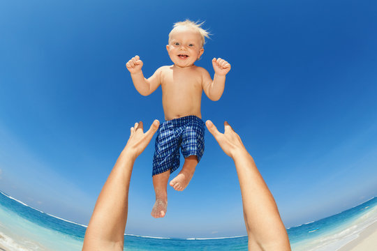 Father Hands Tossing Up The High Air Joyful Baby Boy In On White Sand Sea Beach. Outdoors Healthy Child Activity, Active Lifestyle And Having Fun On Family Summer Vacation With Son On Tropical Island