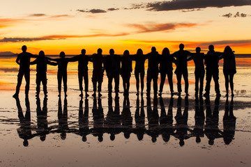Group of friends embraced in the sunset light and wet salt surface in salares de uyuni, Bolivia. Orange sky with clouds background - obrazy, fototapety, plakaty