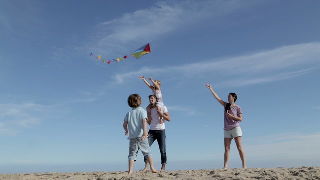 A family of four are playing with a kite. The mother is flying it and the children are chasing after it.