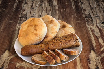 Porcelain Plate with three Pita Bread loafs and Baguette Integral Brown Bread cut in slices, on old Wooden Table. 