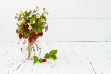 Bouquet of wild strawberries in a glass jug on white old wooden background