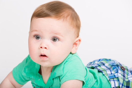 8 month year old baby lays on his stomach looking to the left surprised expression. dressed in a cute green polo shirt and blue plaid shorts.