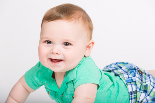 8 month year old baby lays on his stomach looking towards camerawith big smile. dressed in a cute green polo shirt and blue plaid shorts.