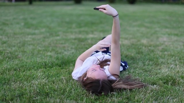 Young beautiful girl lying on the green grass and uses smartphone makes texting and selfie.

