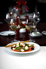 Salad with tomato, mozzarella cheese, mint and olives, beautifully set with glasses of wine in the background