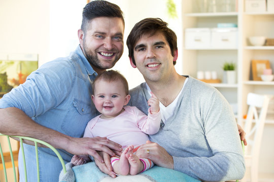 Same sex male couple sitting at home smiling for the camera with their daughter on their lap.