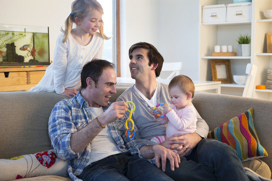 SAme sex male couple sitting in their home and playing with their daughters toys with them.