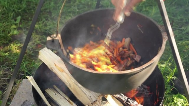 close up view of cooking meat, onion and carrot in a metal pan in a campfire