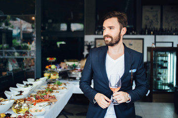 Young elegant man standing in the restaurant, holding a glass of wine. Man's style