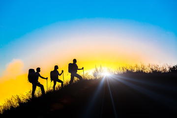 Fototapeta premium People meeting sunrise on team building session Group of people silhouettes walking toward mountain summit with backpacks hiking trekking gear meeting uprising sun sunbeams and blue sky of background
