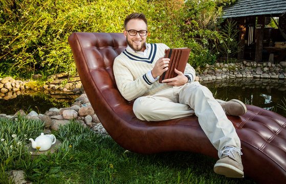 Young Handsome Man In Glasses Sit In Luxury Sofa With IPad In Summer Garden.
