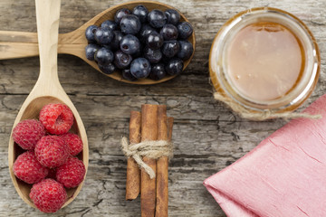 Raspberries and blueberries in spoons with pink napkin and a jar of honey with cinnamon on wooden background. Healthy eating, diet