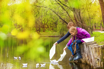 Boy and girl sitting near pond putting paper boats