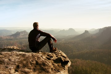 Young man in black sportswear is sitting on cliff's edge and looking to misty valley bellow