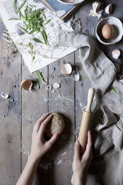 Person handling a dough with a rolling pin over a wooden table with ingredients