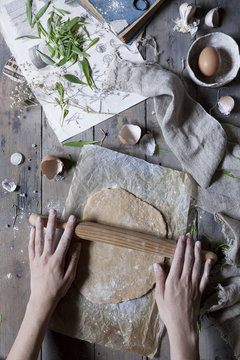 Person handling a dough with a rolling pin over a wooden table with ingredients