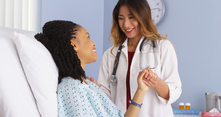 Asian doctor holding hand of black woman patient in hospital bed