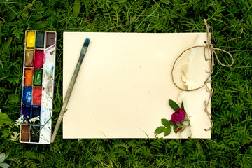 handmade drawing pad with flowers and leaves of wild rose, watercolor paints and brush on the background of grass