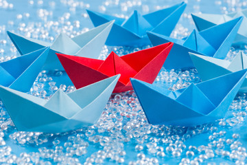 Fleet of blue Origami paper ships on blue waterlike background surrounding a red one