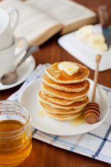 Stack of pancakes with honey syrup and butter in a white plate on a wooden rustic table with book, selective focus