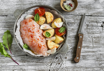 chicken breast, potatoes, onions, tomatoes - raw ingredients for making baked chicken breast with vegetables, vintage pan on a light wooden background