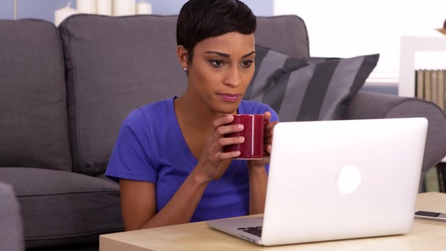 Black woman drinking coffee while reading on laptop
