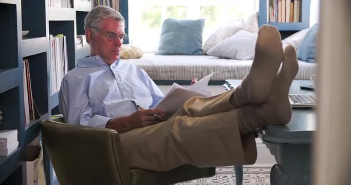 Senior Man In Home Office Doing Paperwork With Feet On Desk