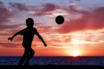 Silhouette of a boy playing football or soccer at the beach with