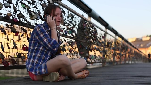 Cute teen girl sitting on the bridge of lovers with snaps, talking on a cell phone.