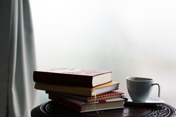 Pile of books lay on the round table and cup against light coming from the window