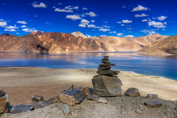 Rocks and reflection of Mountains on Pangong tso (Lake) with blue sky. It is huge lake in Ladakh, It is 134 km long and extends from India to Tibet. Leh, Ladakh, Jammu and Kashmir, India - obrazy, fototapety, plakaty