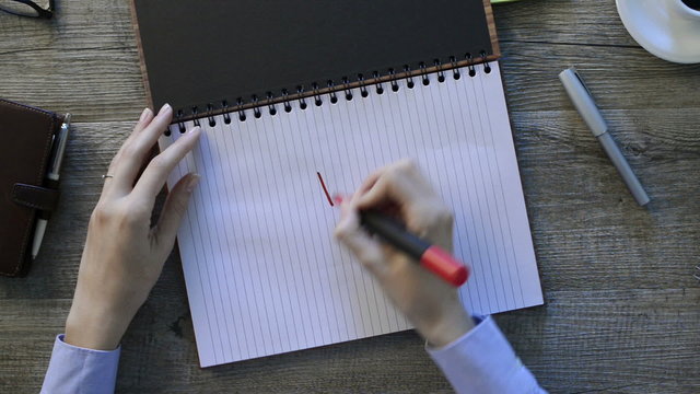 Woman&rsquo;s hand drawing a smiley face on a notebook. Close up of a female hand drawing a smile with red marker pen on a paper in a business desk. Think positive concept.