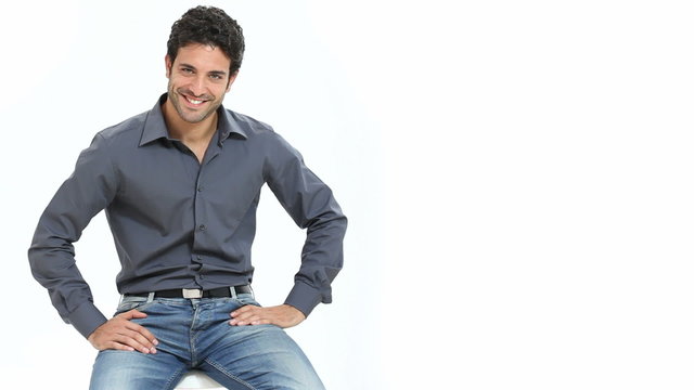 Satisfied and proud young man sitting on chair and looking at camera isolated on white background. Portrait of happy smiling guy with shirt and jeans looking at camera with copy space.