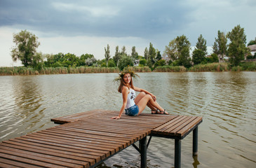 Beautiful young woman with wreath of wildflowers in their hair. Romantic girl with flowers on a wooden pier at the lake. Midsummer.