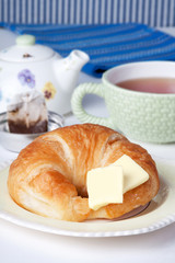 Croissant and Tea – A freshly baked croissant with butter in the foreground. Tea and teapot in background.