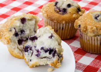 Blueberry Muffin Breakfast – A blueberry muffin cut in half. Whole muffins in the background. On a red checked tablecloth.