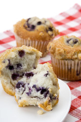 Blueberry Muffins – A blueberry muffin, split in half, on a white plate. Whole muffins in background.
