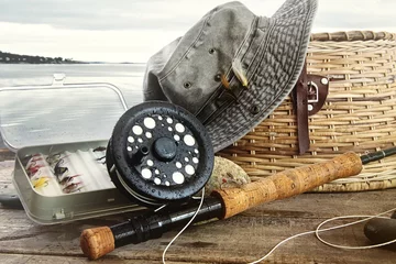 Fototapete Angeln Hut- und Fliegenfischerausrüstung auf Tisch in der Nähe des Wassers