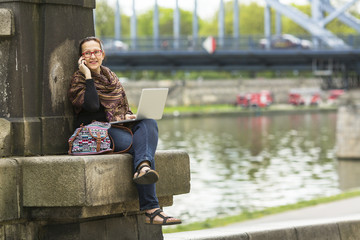 Young woman freelancer sitting on the stone embankment of the river with a laptop and talking on the phone.