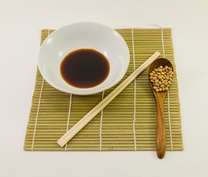 soy sauce in white bowl with bamboo chopsticks and dried soybean in wood spoon on bamboo mat