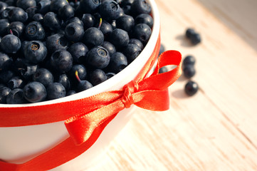 Fresh bilberry in a white ceramic bowl with a red bow on a wooden surface