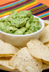 Fresh Guacamole and Chips – A bowl of fresh guacamole. Corn tortilla chips in the foreground. Colorful tablecloth in the background.
