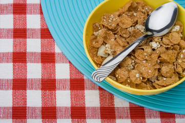 Healthy Bran Cereal – A bowl of bran cereal. Spoon in bowl, blue and red checked background.